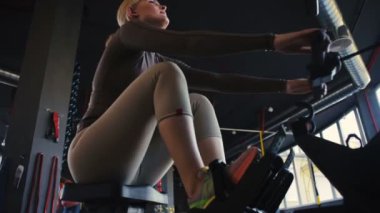Bej renkli spor takım elbiseli profesyonel sarışın sporcu boş spor salonunda modern kürek makinesi üzerinde egzersiz yapıyor.