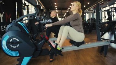 Profesyonel antrenör erkek ve kız kürek makinesinde oturur ve boş modern spor salonunda senkronize bir şekilde kürek çeker.