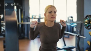 Yoğun, güçlü, genç, sarışın bir kadın bar ve halterli çömelmiş bir şekilde modern spor salonuna yaklaşır.