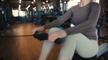 Gri taytlı kaslı kadın, modern spor salonunda arka koltukta sırtüstü oturur, ahşap zemini ağır çekimde kapatır.