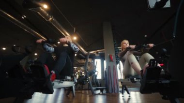 Aktif atletik çift erkek kız spor salonu düşük açılı ağır çekimde modern makinelerde kablolu yaylı sıra yapar.