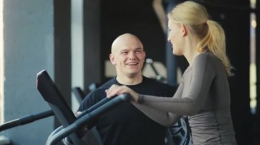 Zayıf aktif kız velosimulator üzerinde egzersiz yapıyor ve modern spor kulübünde ağır çekimde gülümseyen kel koçu dinliyor.