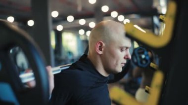 Siyah giysili kel adam halter alır ve bulanık kadın siluetine karşı çömelir.