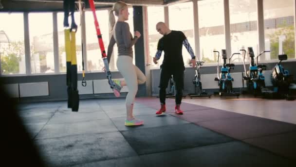 职业太极拳教练在现代体育馆的中等视野中以特殊的健身带控制着蹲着的年轻女子 — 图库视频影像