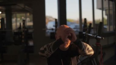 Siyah eşofmanlı güçlü kel adam spor salonundaki siyah makinelere karşı parlak batan güneş ışığı altında zıplıyor.