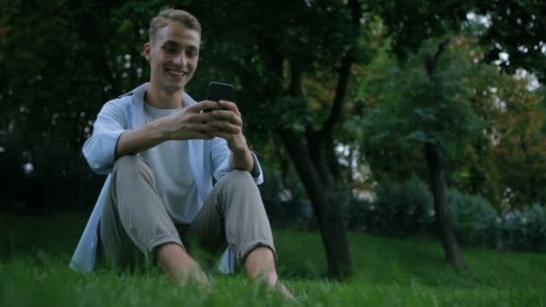 Barefoot guy sits on green park grass and smiles typing Stock Footage