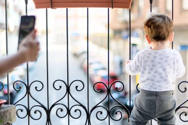 Bir buçuk yaşında bir çocuk balkonda oynuyor ve karantinada geçen arabaları izliyor çünkü dışarı çıkamıyor.