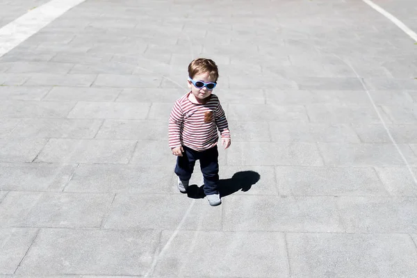A year and a half old boy walking down the street with blue sunglasses on