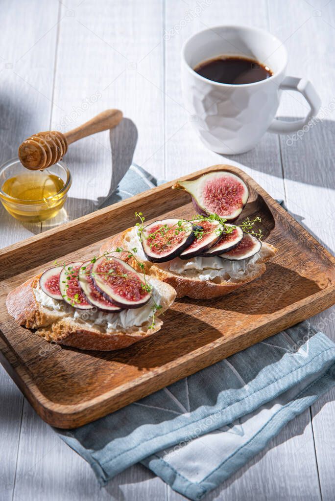 Breakfast with fig, cheese and honey sandwich on a wooden board and cup of coffee on white table, sunny morning