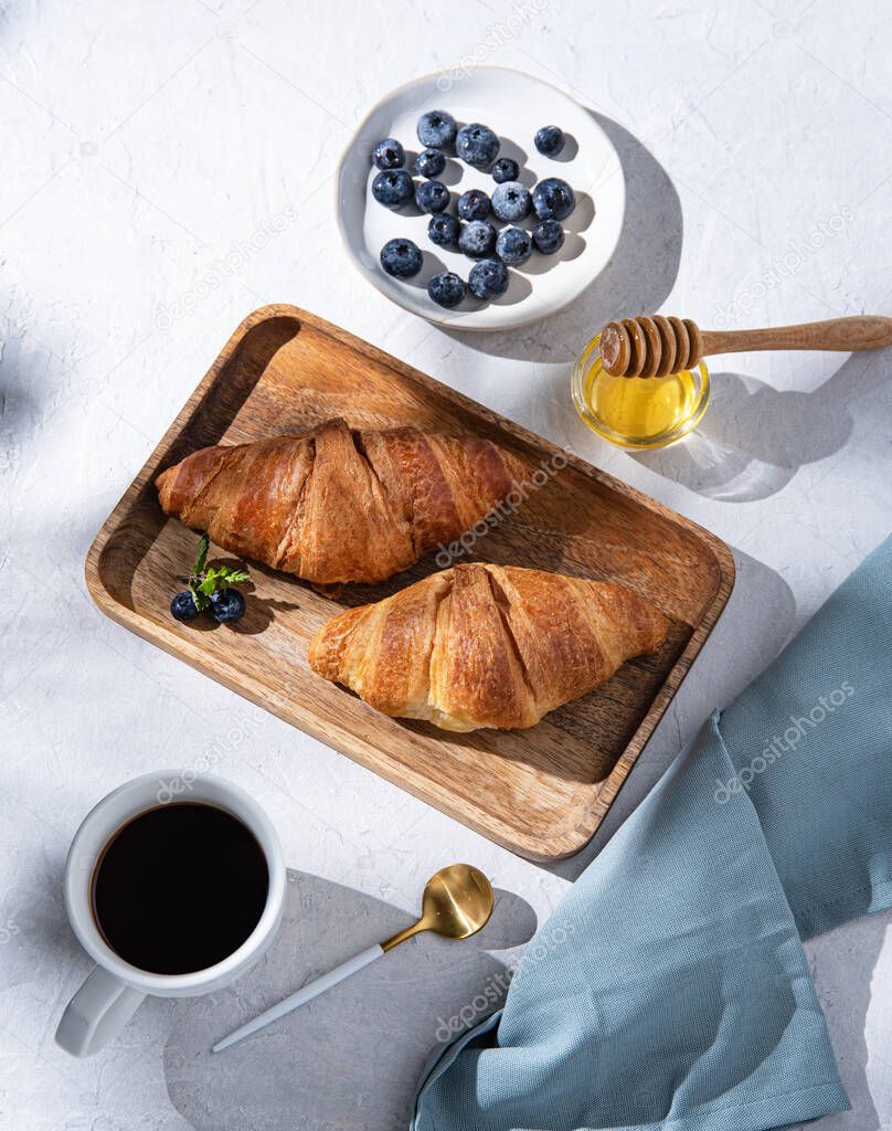 two  croissants on a cutting board  with cup of coffee, honey and  blueberries  on white background. flat lay and top view