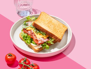 Pastırmalı, peynirli, domatesli, marullu, roka soslu ve pembe arka planda bir tabakta bir tost ekmeği, bir bardak suyla. Menü kafenin reklam afişi için sağlıklı ve besleyici kahvaltı konsepti. Boşluğu kopyala.