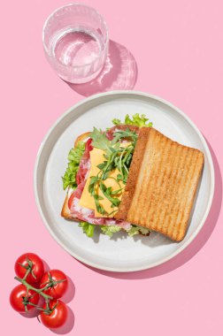 Pastırmalı, peynirli, domatesli, marullu, roka soslu ve pembe arka planda bir tabakta bir tost ekmeği, bir bardak suyla. Menü kafenin reklam afişi için sağlıklı ve besleyici kahvaltı konsepti. Üst görünüm.