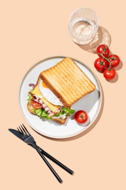 Yumurtalı, pastırmalı, peynirli, domatesli, salatalı ve soslu sandviç bej bir arka planda, bir bardak su ve çatal bıçaklı. Reklam menüsü ya da afiş kafe için sağlıklı ve besleyici tost kahvaltısı konsepti. Üst görünüm.