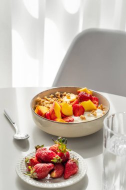 Doğal Yunan yoğurdu, çilek ve şeftalili granola ve müsli bir bardak su, taze böğürtlen, sandalye ve sabah güneşiyle birlikte beyaz bir masa üzerinde beyaz bir tabakta. Sağlıklı ve besleyici kahvaltı konsepti. Boşluğu kopyala.