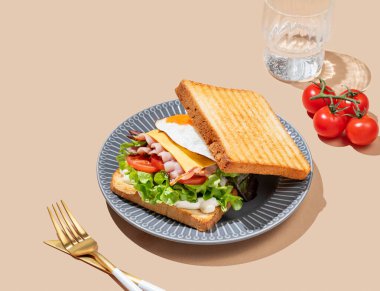 Yumurtalı, pastırmalı, peynirli, domatesli, salatalı ve soslu sandviç bej bir arka planda, bir bardak su ve çatal bıçaklı. Reklam menüsü ya da afiş kafe için sağlıklı ve besleyici tost kahvaltısı konsepti. Boşluğu kopyala.