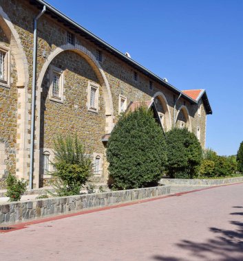 Kemerli camları ve yeşil çalıları olan taştan bir bina, gün ışığında, asfalt bir yolda. Tarihi mimari ve kültürel miras alanı kavramı. Kıbrıs.