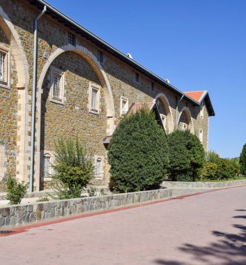 Kemerli camları ve yeşil çalıları olan taştan bir bina, gün ışığında, asfalt bir yolda. Tarihi mimari ve kültürel miras alanı kavramı. Kıbrıs.