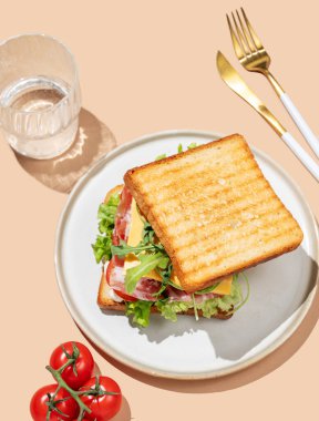 Pastırmalı, peynirli, domatesli, marullu, roka soslu, parlak bej arkaplanlı ve bir bardak suyla servis edilmiş bir tost ekmeği. Menü kafenin reklam afişi için sağlıklı ve besleyici kahvaltı konsepti. Üst görünüm.