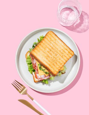 Pastırmalı, peynirli, domatesli, marullu, roka soslu ve pembe arka planda bir tabakta bir tost ekmeği, bir bardak suyla. Menü kafenin reklam afişi için sağlıklı ve besleyici kahvaltı konsepti. Üst görünüm.