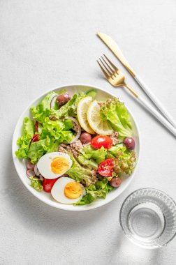 Kiraz domatesli ton balığı salatası, yumurta, zeytin ve salatalık beyaz bir tabakta, arka planda bir bardak su ve çatal bıçakla. Sağlıklı Akdeniz yemekleri konsepti. Üst görünüm, alanı kopyala.