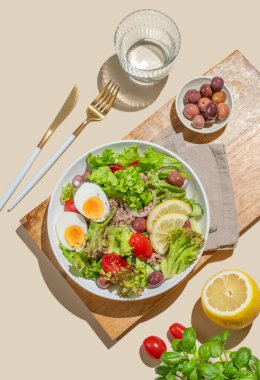 Kiraz domatesli ton balığı salatası, yumurta, zeytin ve salatalıklar tahta bir tabakta, parlak sarı bir zeminde, bir bardak suyla. Reklam menüsü kafesi için sağlıklı Akdeniz yemekleri konsepti. Üst görünüm, alanı kopyala.