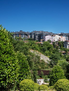 Ormanlık bir yamaçtaki kentsel yerleşim alanı, açık mavi gökyüzü, doğa ve modern yaşam alanları arasındaki zıtlığı gösteriyor. İstanbul, Türkiye.