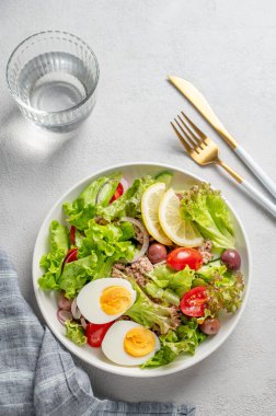 Kiraz domatesli ton balığı salatası, yumurta, zeytin ve salatalık beyaz bir tabakta, arka planda bir bardak su, peçete ve çatal bıçakla. Sağlıklı Akdeniz yemekleri konsepti. Üst görünüm, alanı kopyala.