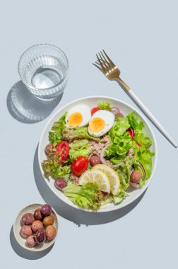 Mavi bir zemin üzerinde mavi bir tabakta bir bardak su ve çatal bıçakla kiraz domatesli ton balığı salatası, yumurta, zeytin ve salatalık. Reklam menüsü kafesi için sağlıklı Akdeniz yemekleri konsepti. Üst görünüm, alanı kopyala.