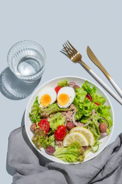 Mavi bir zemin üzerinde mavi bir tabakta, kiraz domatesli, yumurtalı, zeytinli ve salatalıklı ton balığı salatası ve bir bardak su, peçete ve çatal bıçak. Reklam menüsü kafesi için sağlıklı Akdeniz yemekleri konsepti. Üst görünüm, alanı kopyala.