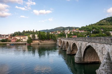 Eski bir taş köprü, sakin bir nehri güzel bir kasabayla geçer ve parlak mavi bir gökyüzünün altında yumuşak bulutlarla yemyeşil tepeler oluşturur. Drine Köprüsü. Visegrad, Bosna-Hersek.
