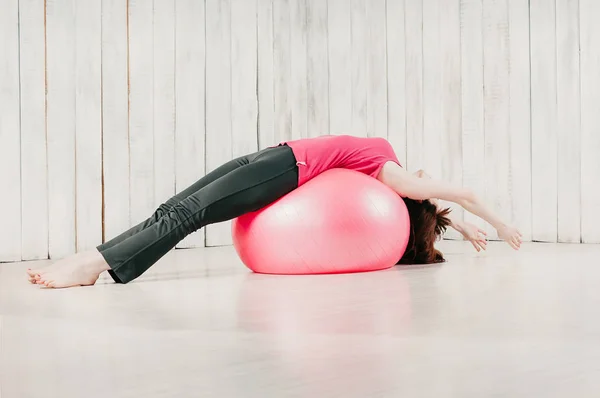 a young pretty woman in a pink top, lying on a pink fitball in a gym, holding  balance, light floor and background