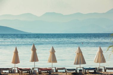 Beyaz plaj şemsiyeleri, mavi deniz ve büyük Puslu Dağlar ufukta, güneşli bir gün
