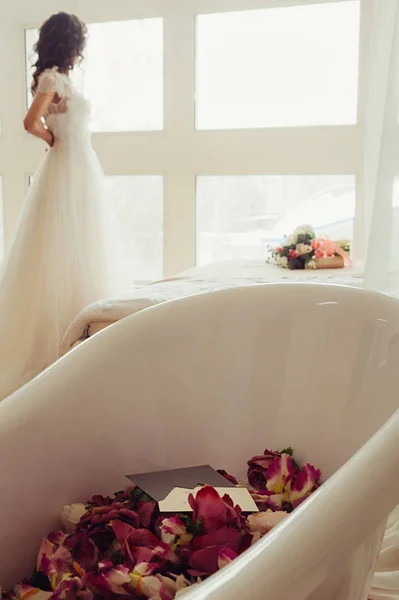 A white bath with red flowers and a letter in the envelope, with blurred bride buttoning her dress in the background looking into window, her wedding bouquet on a white bed next to her