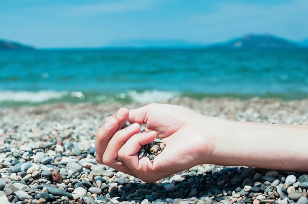 Closeup view of a hand holding pebbles on a beach, turquoise sea water in the background. Summer holiday concept