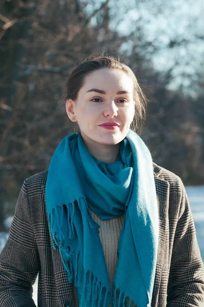 Street style Fashion portrait cute young woman in trendy casual clothes, in a snowy park. Brown overcoat, blue scarf. Sunny winter day, city lifestyle. Classic look