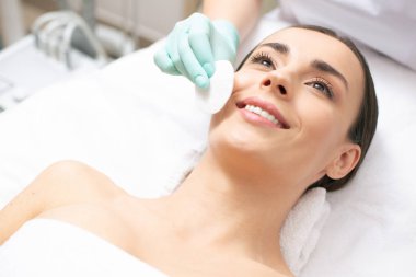 Beyaz havlu ve kozmetolojist üzerinde yatan doğal makyaj ile gülümseyen kadın cildini temizlerken bir pamuk ped kullanarak