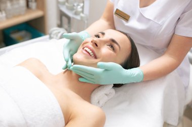 Tıbbi kanepede yatan ve gülümseyen mutlu genç bayan Yakın profesyonel kozmetolojist dikkatle yüzüne dokunarak iken