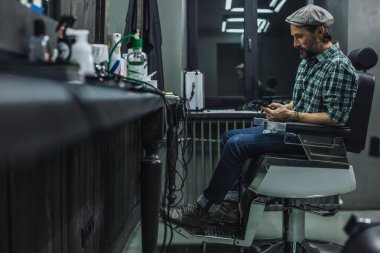 Ayna önünde berber koltuğunda oturan ve onun modern akıllı telefon ekranına bakarak düz kap çekici adam tam uzunlukta