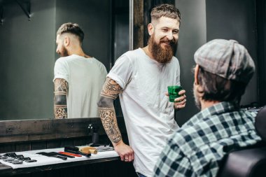 Relaxed bearded barber standing with a mirror behind him and smiling to the visitor while holding a glass