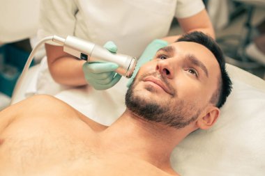 Klinikte yatan ve oksijen meso tedavisi yürüten kozmetolojist gülümseyen sakallı bir adamın yakın fotoğrafı