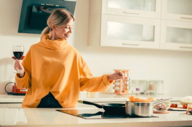 Neşeli genç bayan bir kadeh şarap ile mutfakta ayakta ve kızartma tavada yemeğe tuz eklerken gülümseyerek