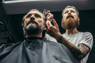 Sakin sakallı adam oturan ve profesyonel berber dikkatle saçlarını kesme sırasında seyir