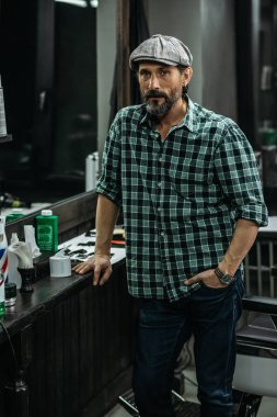 Sakin çekici sakallı adam damalı gömlek berber dükkanı tezgahında bir el ve cebinde bir el ile ayakta