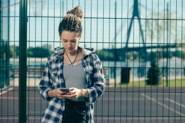 Düşünceli genç bayan zincir bağlantı çit yanında duran ve onun akıllı telefon ekranına bakarak