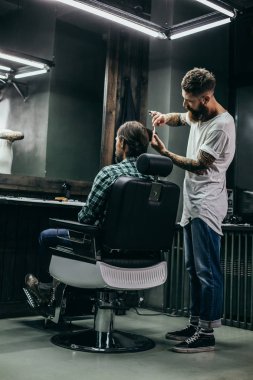 Bearded barber in casual clothes standing next to his client and looking concentrated while combing his hair