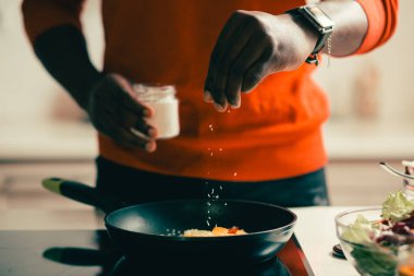 Bileğinde akıllı saat olan adam hindistan cevizi gevreği bir kavanoz tutan ve kızartma tavada yemek için küçük bir miktar ekleyerek