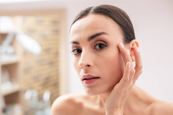 Close up photo of attentive young woman with natural makeup standing in front of the camera and thoughtfully touching skin near the eye