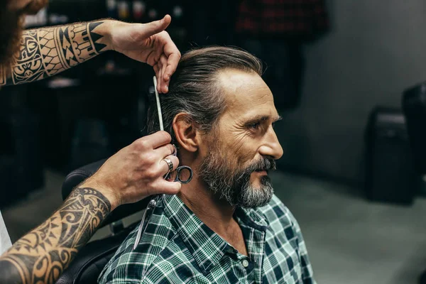 Calm handsome man sitting and smiling while professional barber standing next to him and doing haircut