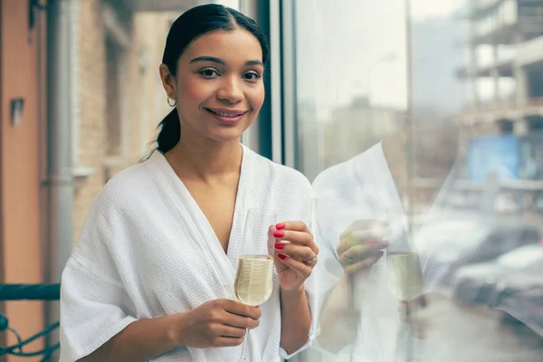Young relaxed woman in a white bathrobe standing near the window alone and smiling with a glass of champagne in her hands