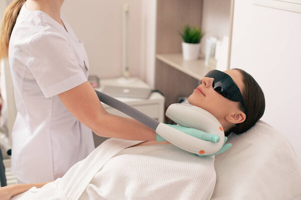 Young woman lying on the medical couch with black protective glasses on her face and smiling while having laser hair removal procedure